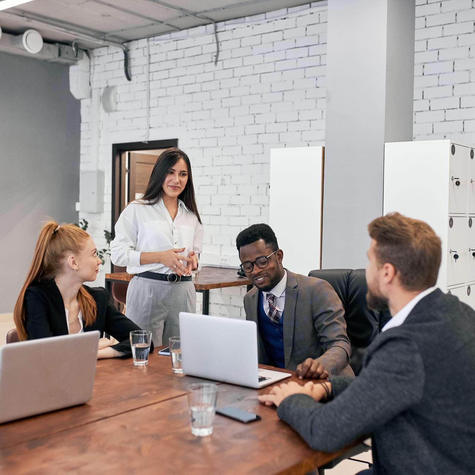 Four people in a modern office, three seated with laptops, one standing, engaged in a discussion. White brick walls and wooden table visible.