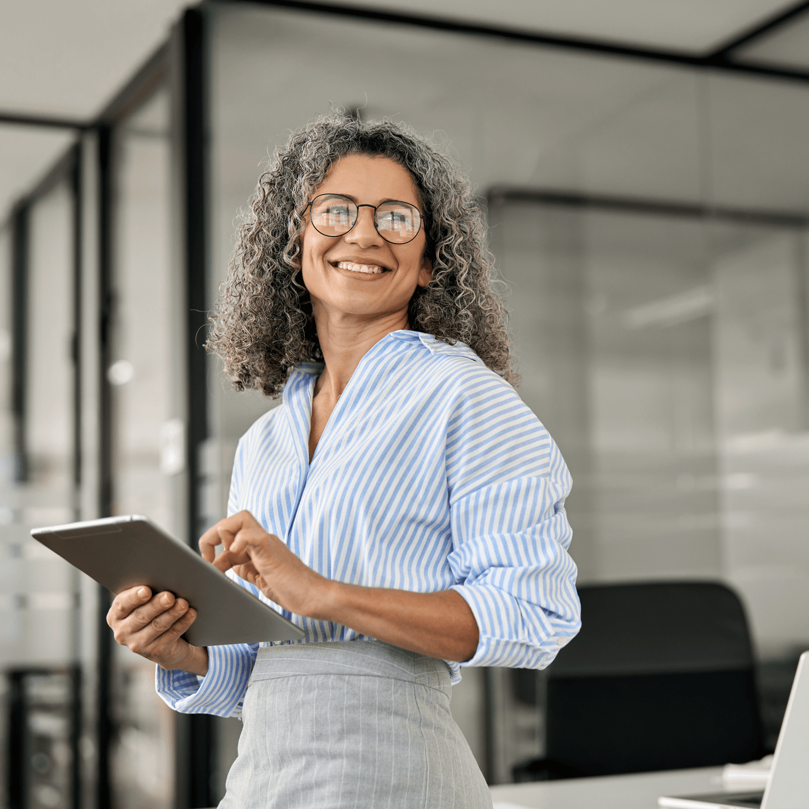 Smiling woman with curly hair and glasses holds a tablet in a modern office setting, wearing a striped shirt and gray skirt.