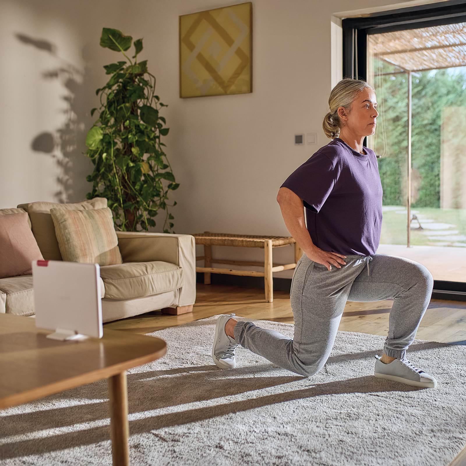 A woman in a purple shirt and gray pants performs a lunge exercise in a cozy living room with a large plant and a window view.