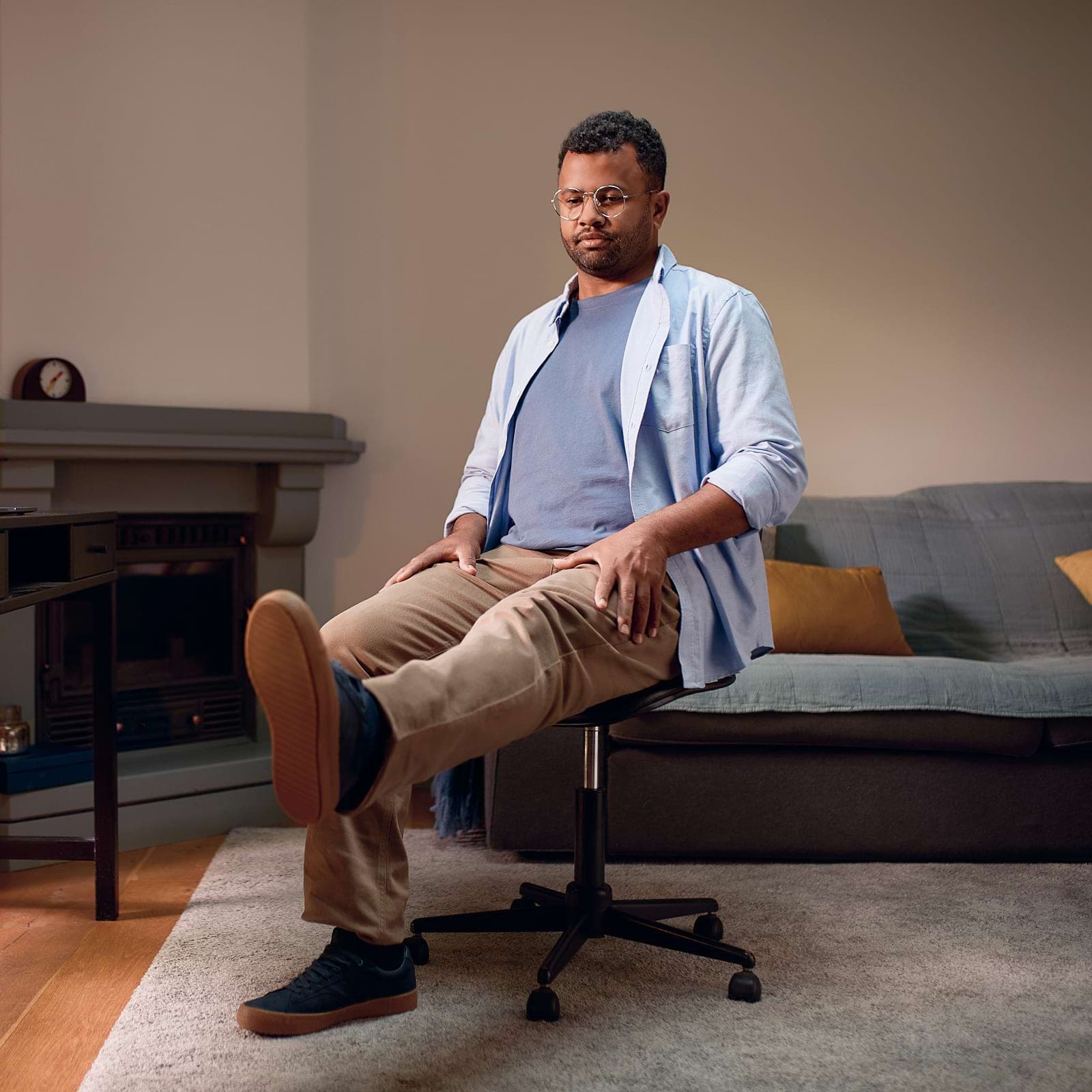 Man sitting on a swivel chair in a living room, wearing a blue shirt and glasses, with a neutral expression.