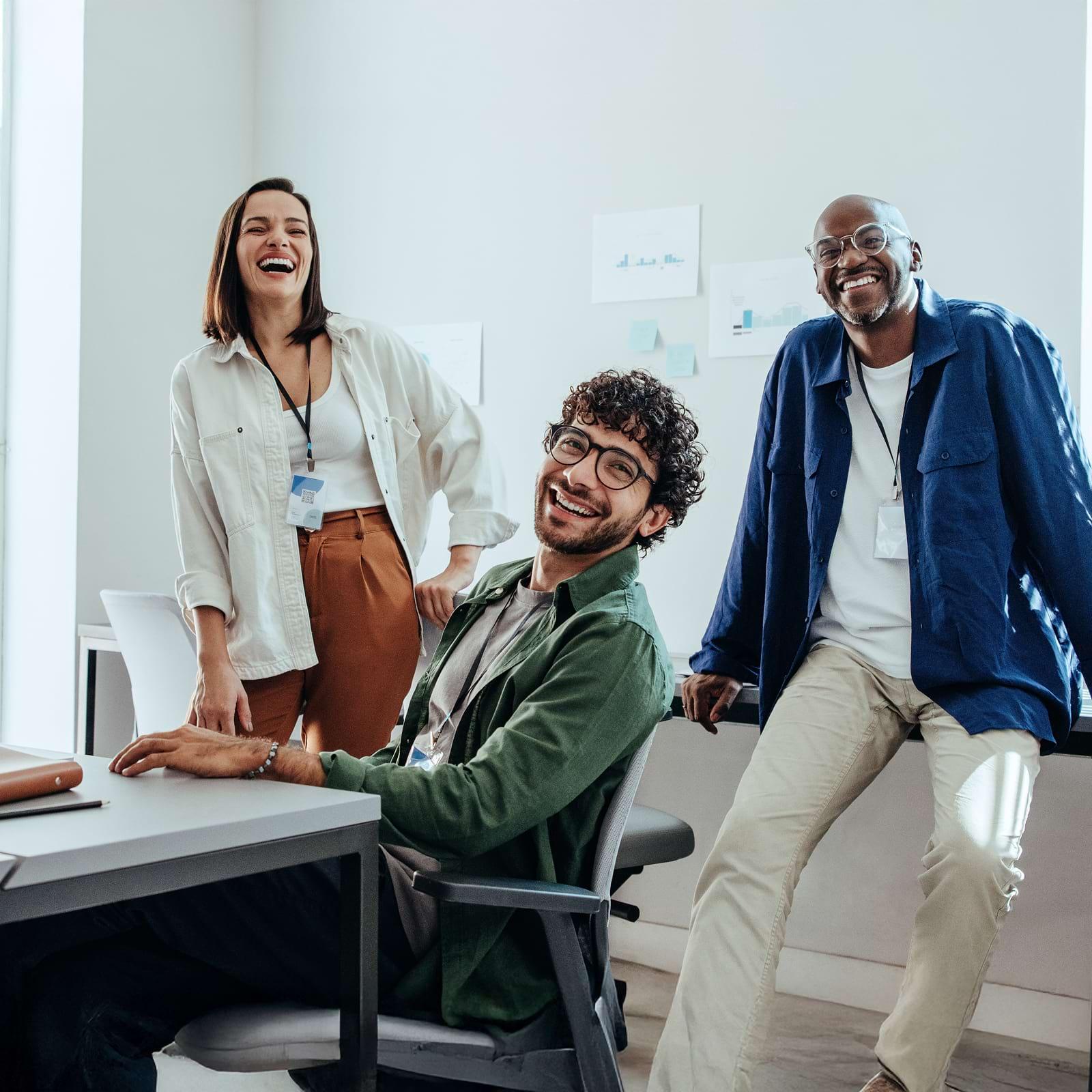 Three people in a bright office, smiling and laughing. One is seated at a desk, and two are standing, all wearing casual business attire.