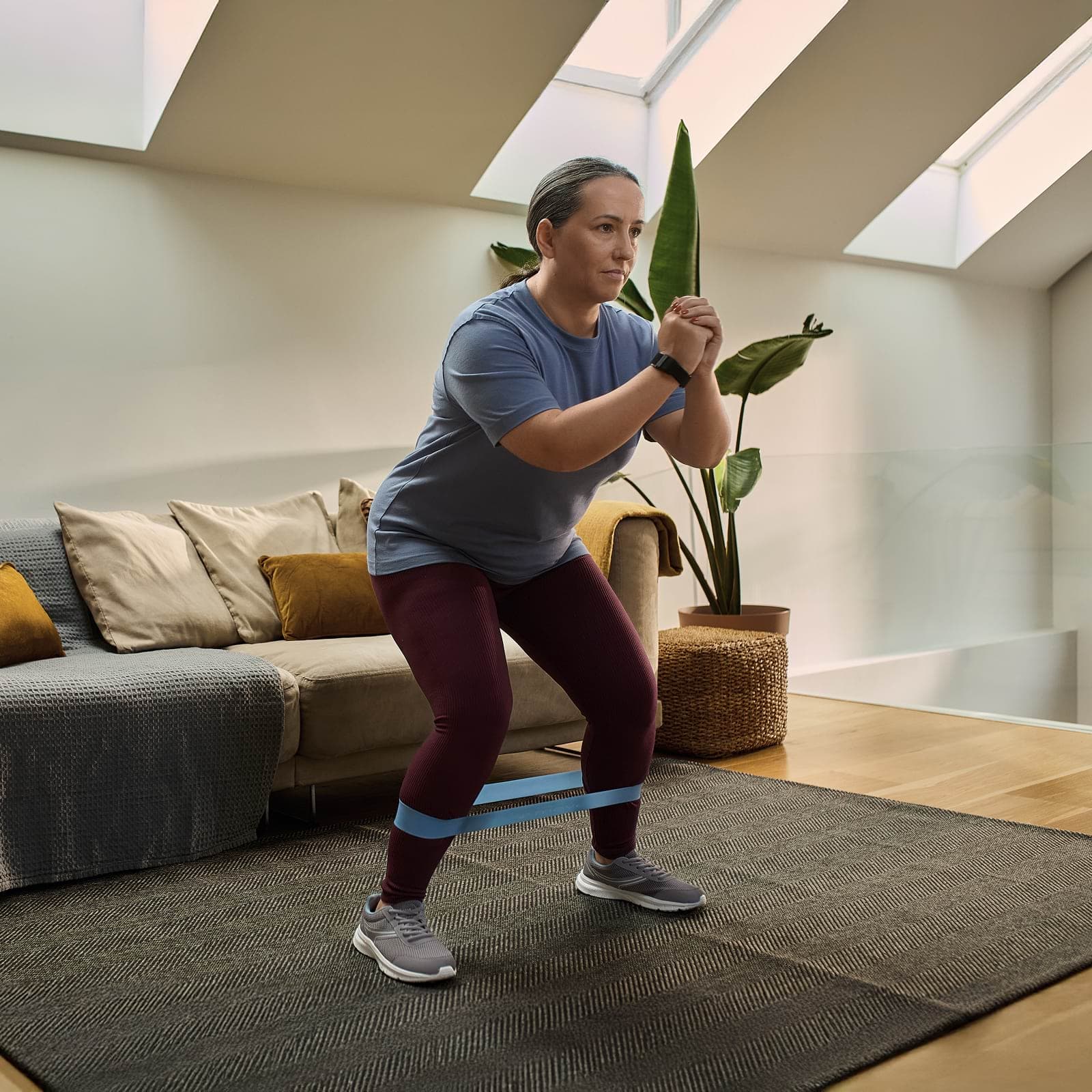 A person exercises in a living room, wearing a resistance band around their legs, with a sofa and plants in the background.