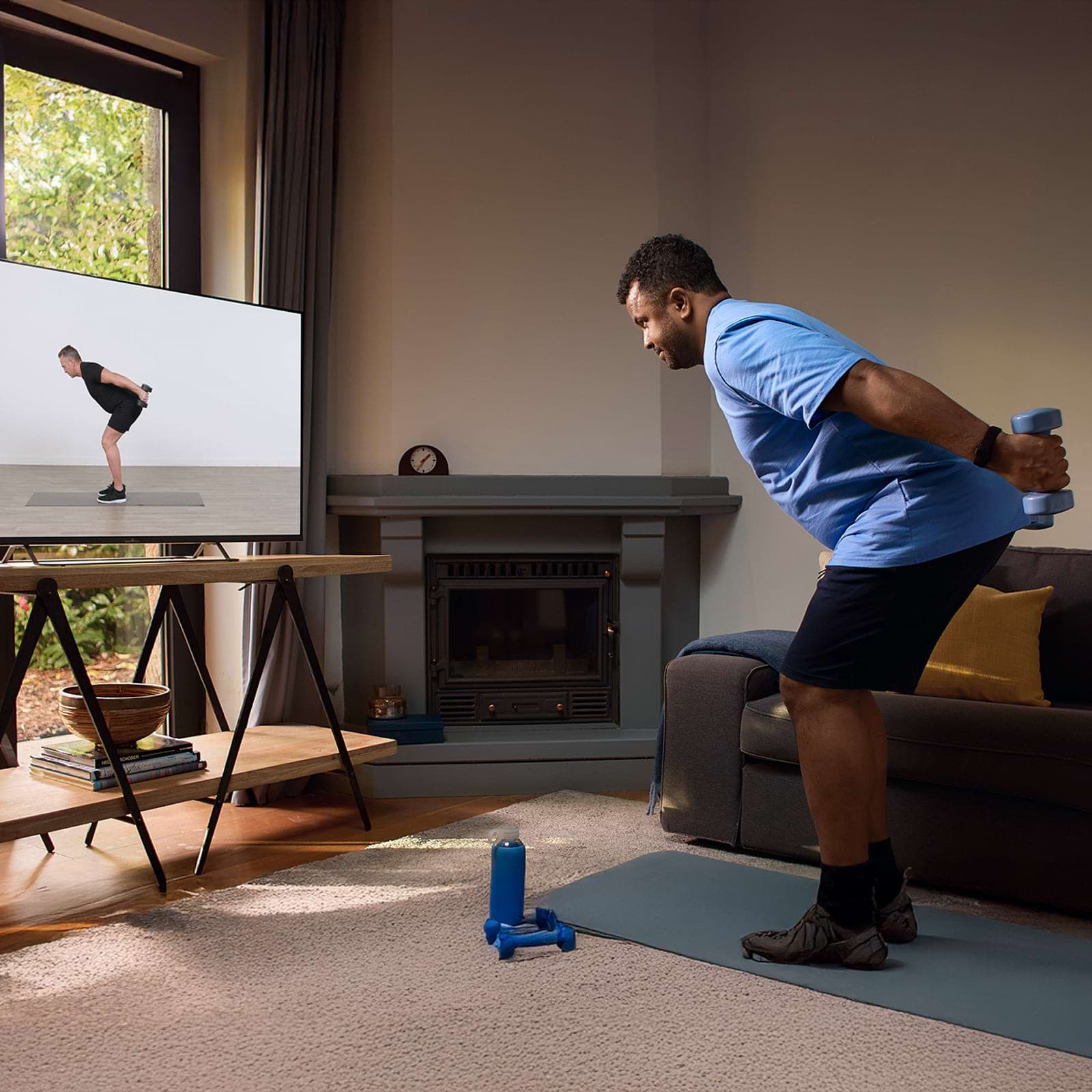 A man exercises at home, following a workout video on TV, holding dumbbells and bending forward. A mat and additional weights are on the floor.