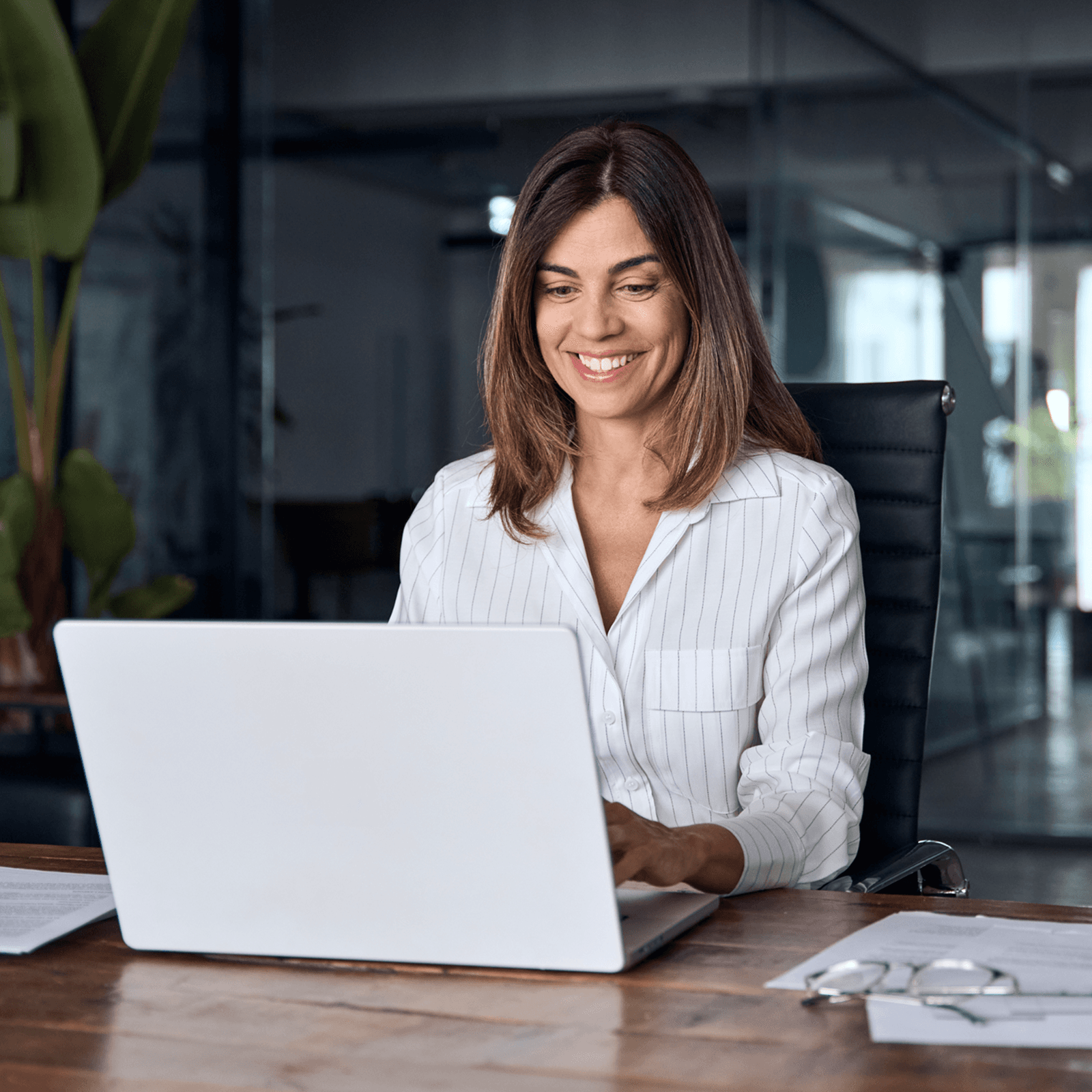 Smiling woman in a white shirt working on a laptop at a wooden desk in a modern office with glass walls and plants.