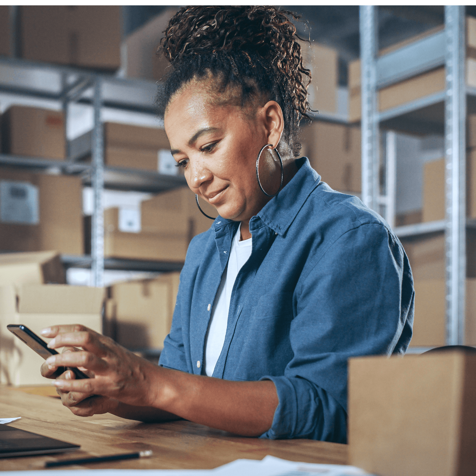 Woman in a warehouse checking her phone, surrounded by boxes and shelves, wearing a blue shirt and hoop earrings, seated at a wooden table.