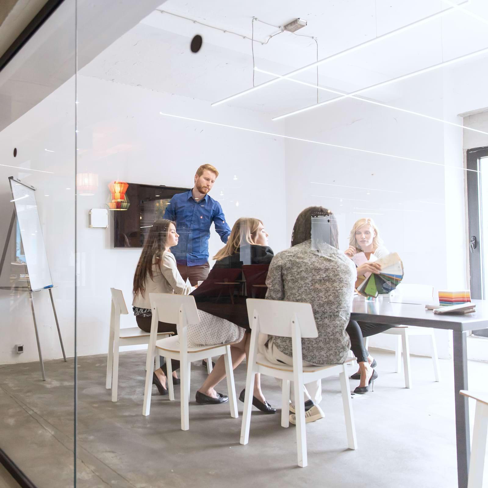 People in a modern glass-walled office, engaged in a meeting around a table with a flip chart and a wall-mounted screen.