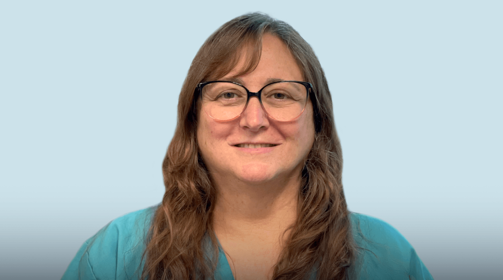 Smiling person with long hair and glasses, wearing a teal top, against a light blue background.