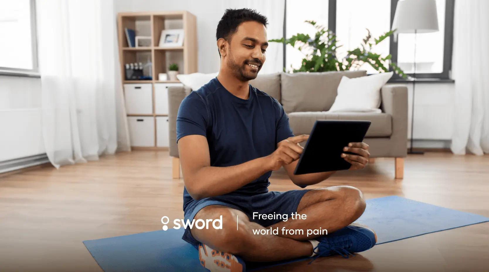 Man sitting cross-legged on a yoga mat in a living room, using a tablet. Text reads "Sword: Freeing the world from pain."