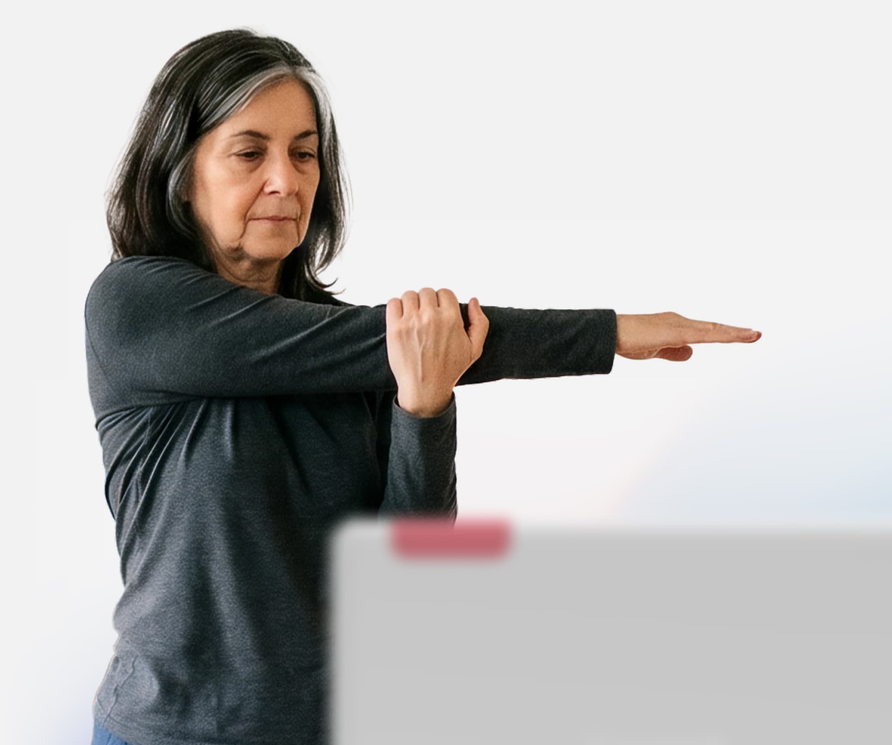 A woman with gray-streaked hair stretches her left arm across her chest, using her right hand to hold it, while standing indoors and looking focused.