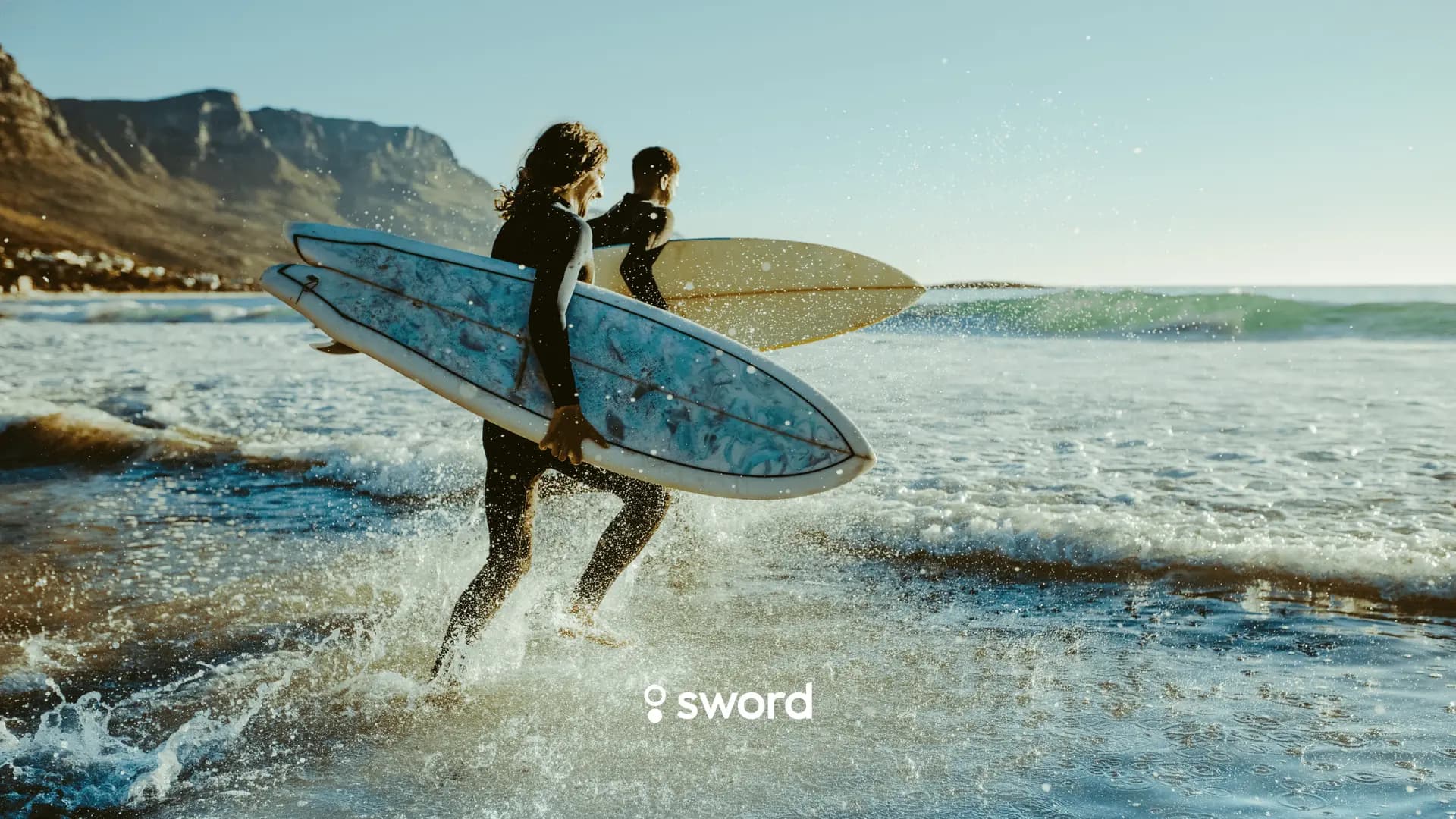 Two surfers in wetsuits carry surfboards into the ocean, with waves and mountains in the background.