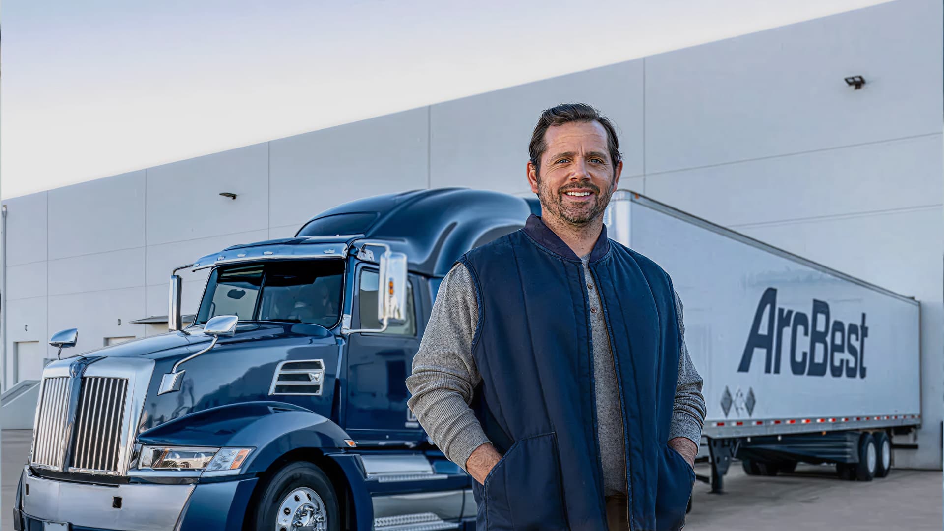 Smiling man standing in front of a blue semi truck and white ArcBest trailer outside a warehouse.