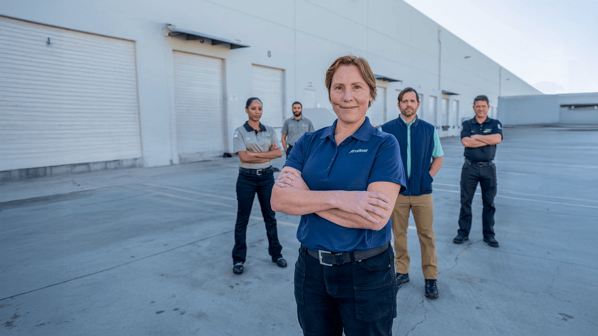 Five warehouse workers stand outside a loading-dock warehouse, arms crossed, with a woman in a navy polo at the front.