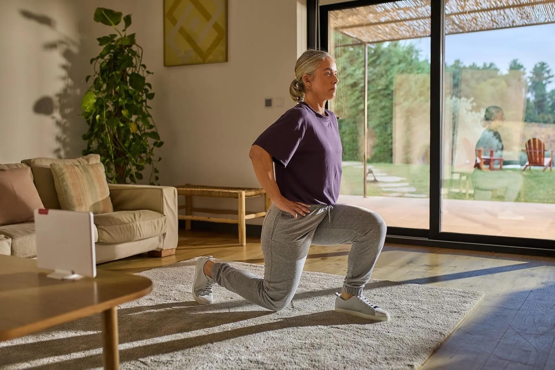 Woman doing lunges in a living room, wearing a purple shirt and gray pants, with large windows showing a garden view.
