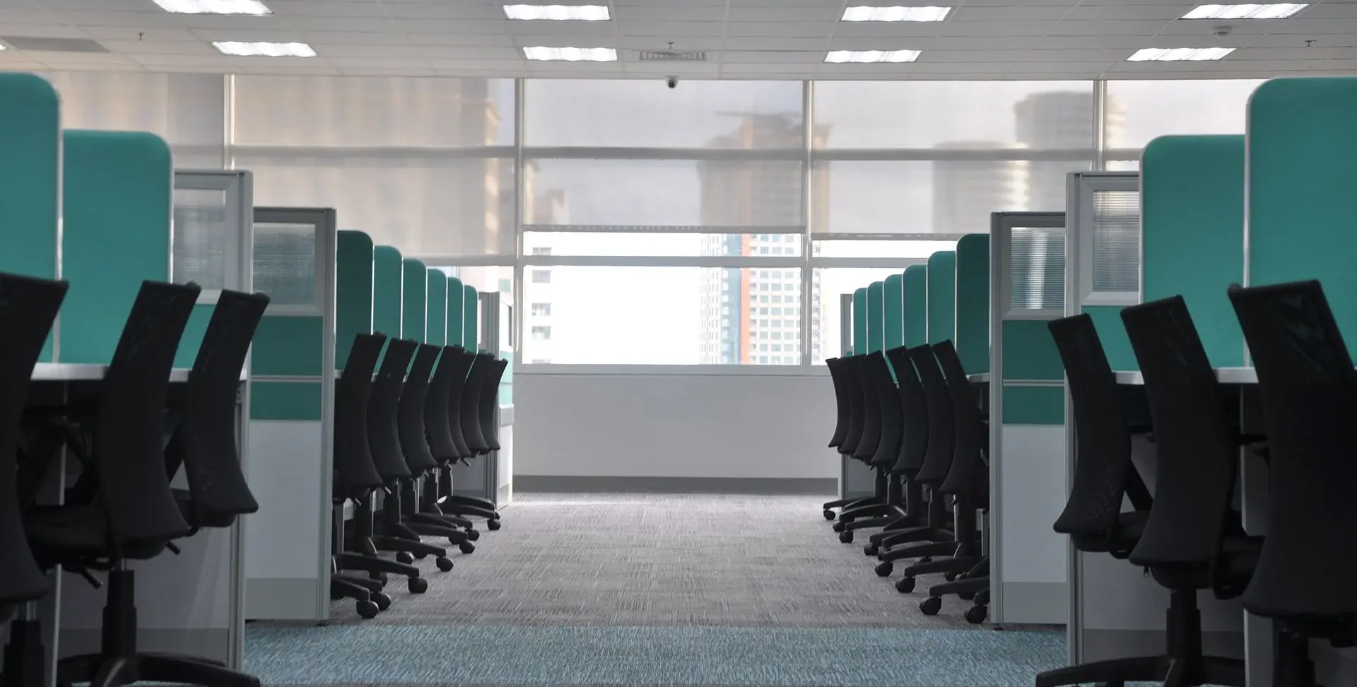 A modern office space with rows of empty black chairs and teal dividers, facing large windows with a cityscape view.