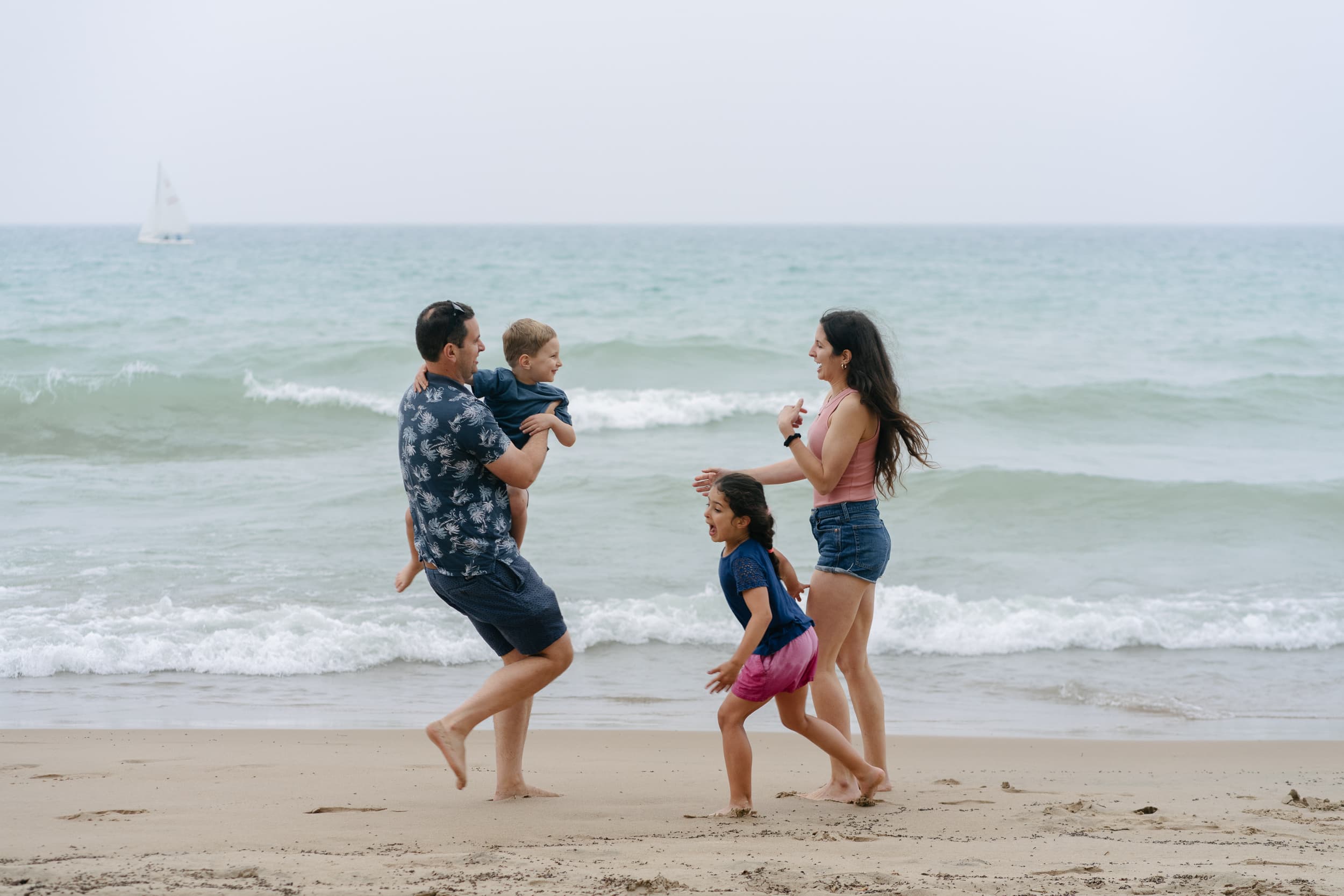 A family playing joyfully on a sandy beach near the ocean. A man carries a child, while a woman and a girl run nearby. A sailboat is in the distance.