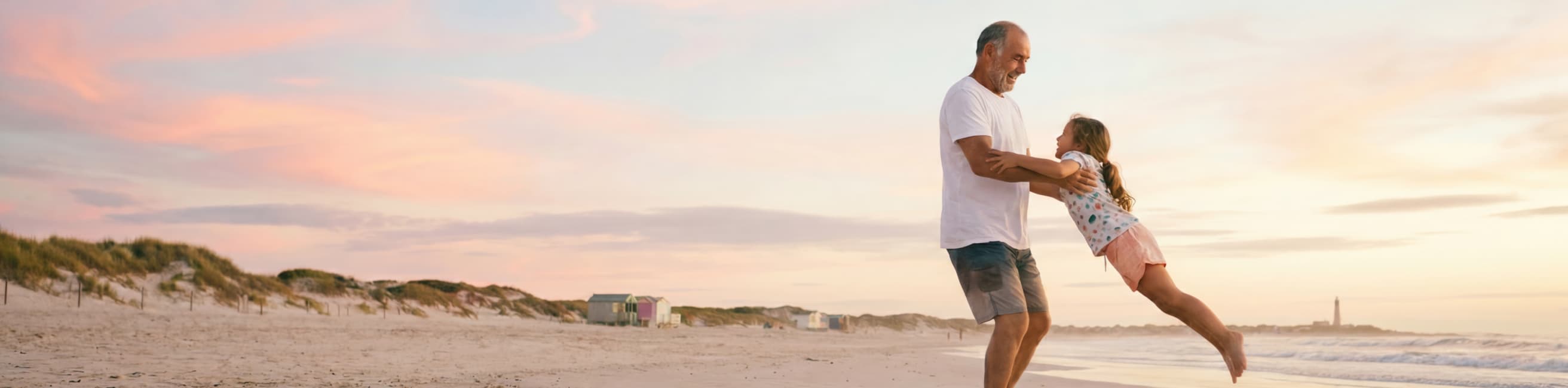 A man spins a smiling young girl by the arms on a sandy beach at sunset, with pastel clouds in the sky and gentle waves in the background.
