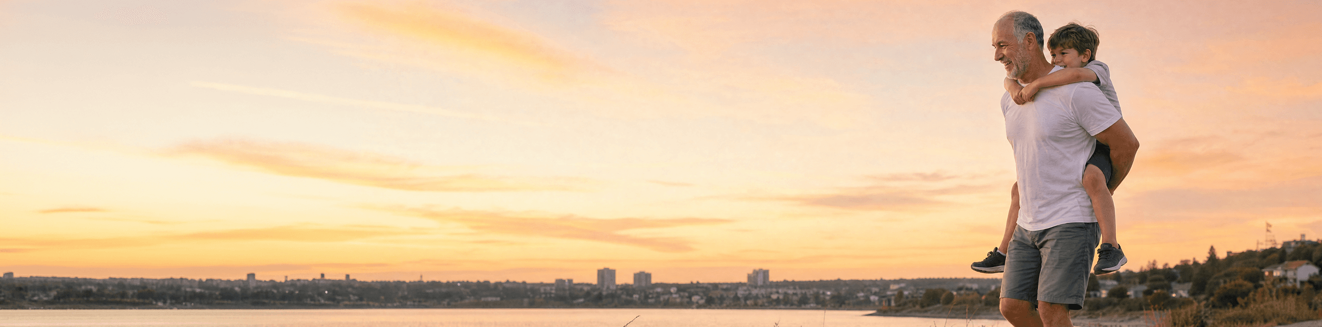 An older man carries a young boy on his back by a lake at sunset, both smiling and enjoying the scenic view. City buildings are visible in the distance under a colorful sky.