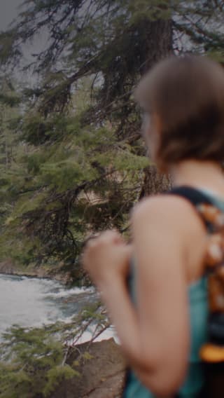 A person with short hair and a backpack stands near a river, looking at the flowing water, with tall evergreen trees in the background.