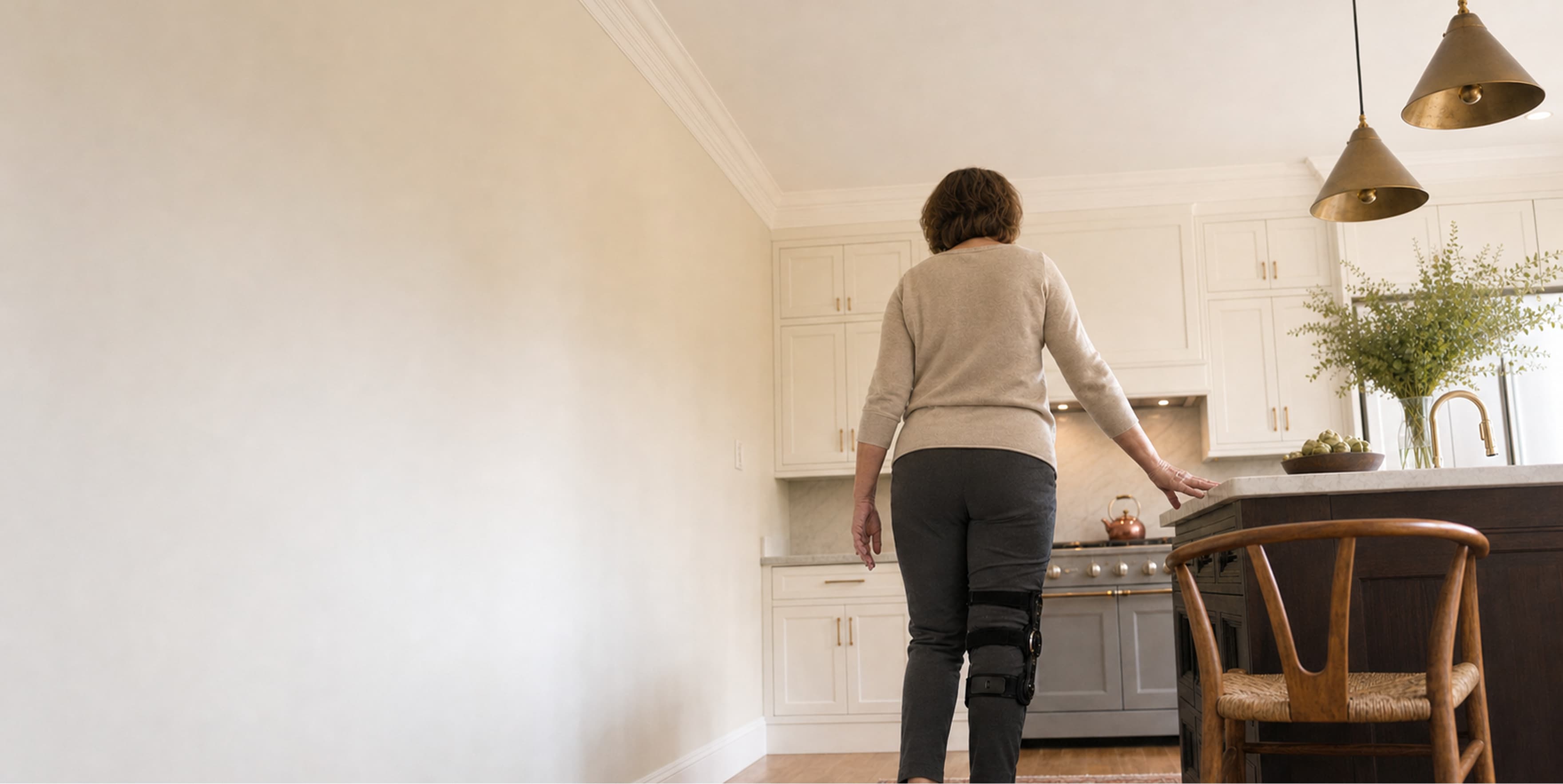 A person with a knee brace stands in a modern kitchen, facing away and lightly touching the counter. The kitchen features white cabinets, pendant lights, and a wooden chair.