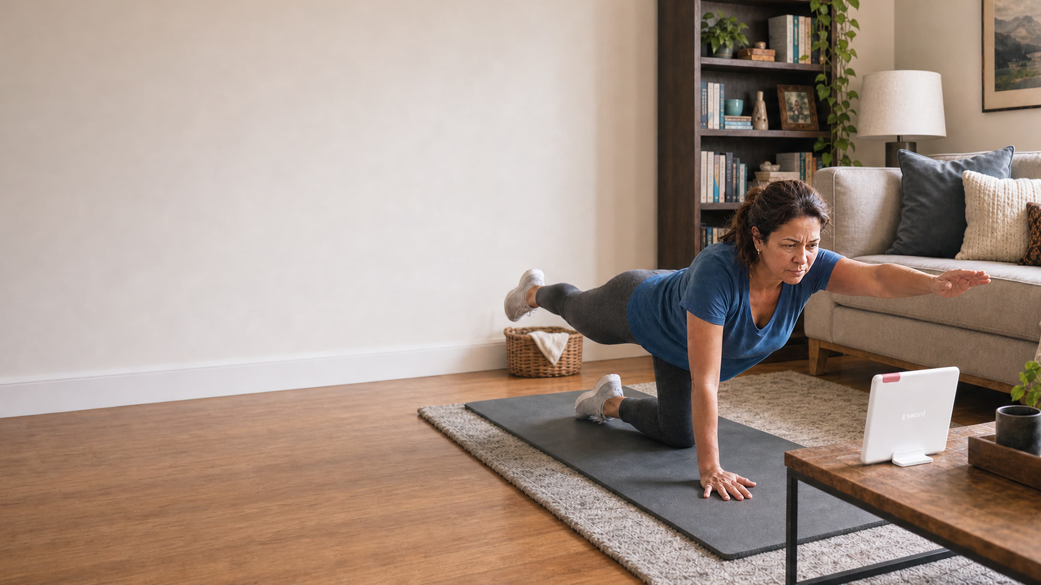 A woman exercises on a yoga mat in her living room, balancing on one hand and one knee while looking at a tablet on a stand. Shelves, a lamp, and a sofa are in the background.