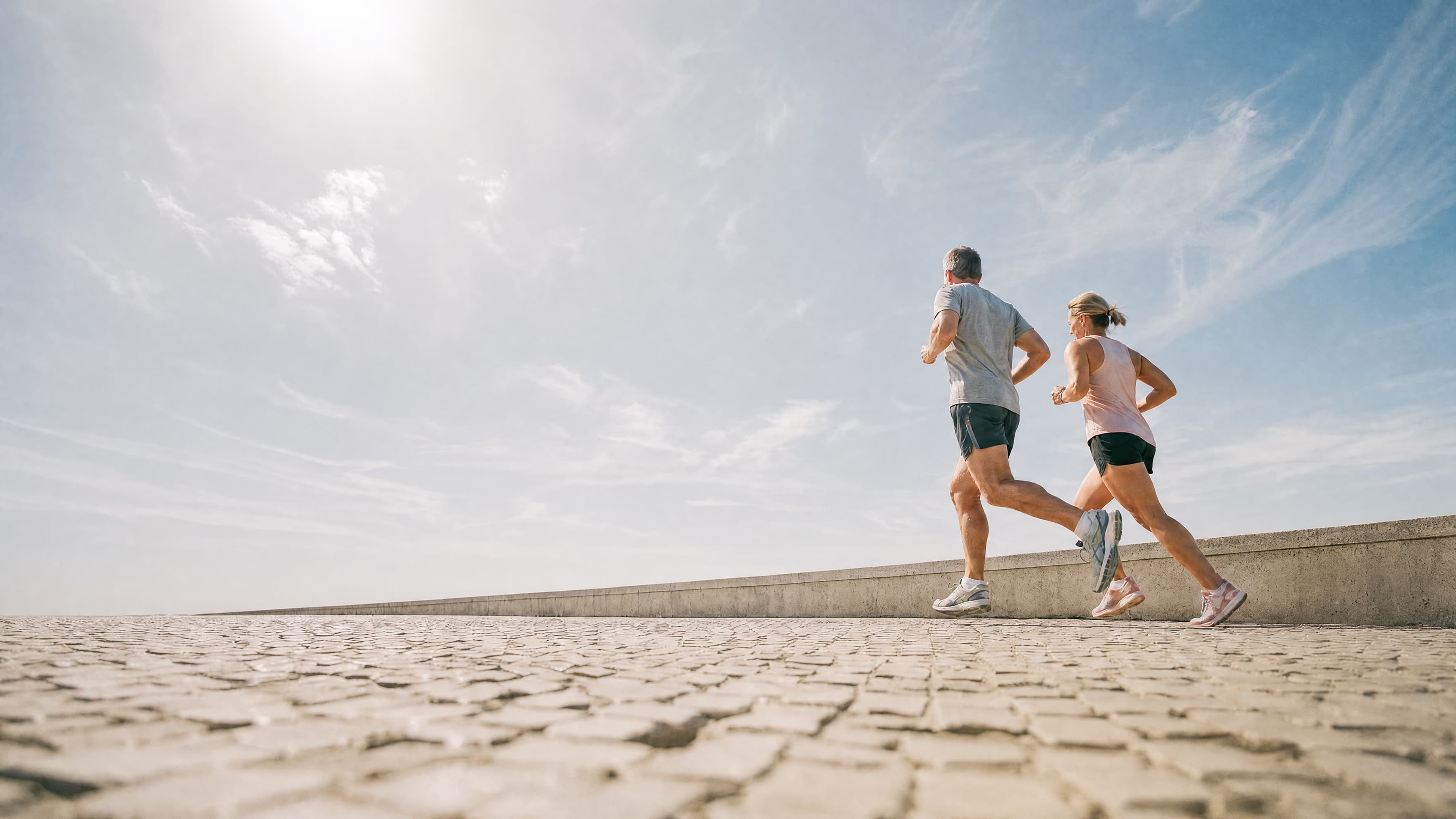 A man and a woman jog together on a sunlit stone pathway under a clear, blue sky with wispy clouds. The bright sun shines above, casting their shadows on the ground.