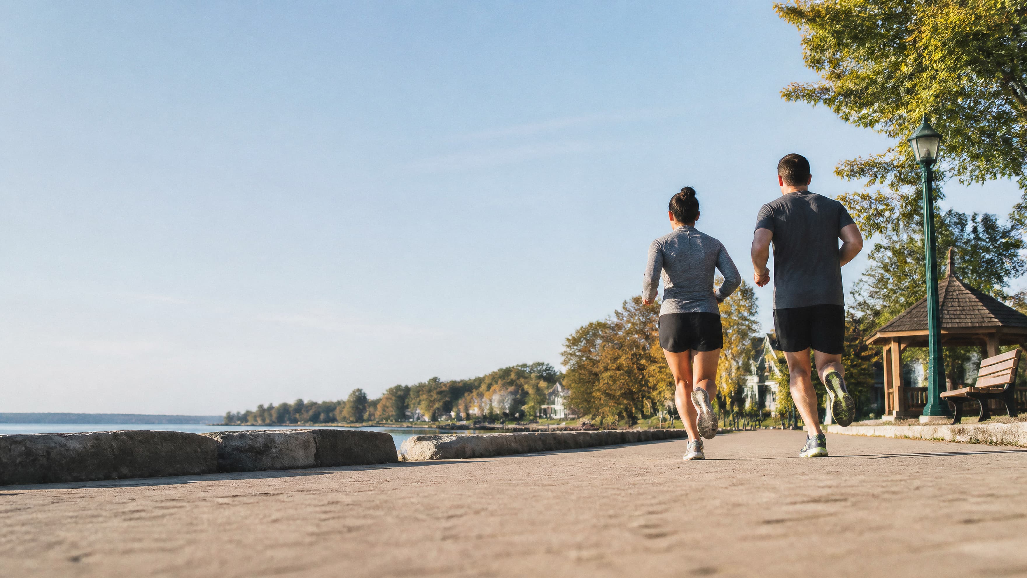 A woman and man jogging away from the camera on a paved waterfront path on a sunny day, with trees, a gazebo, and a calm lake in the background.