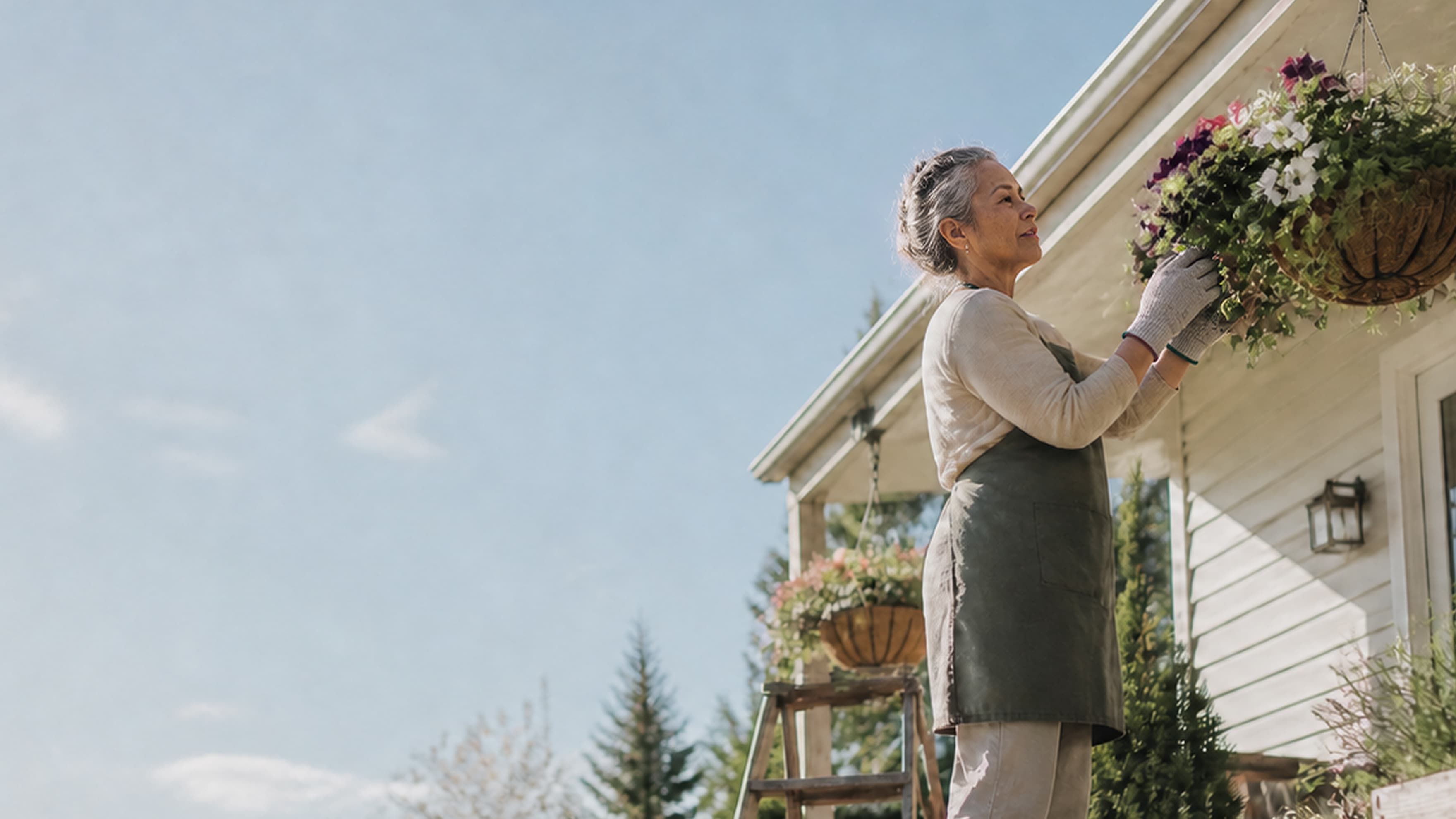 An older woman wearing gloves and an apron tends to hanging flower baskets outside a house on a sunny day, with trees and a ladder nearby.