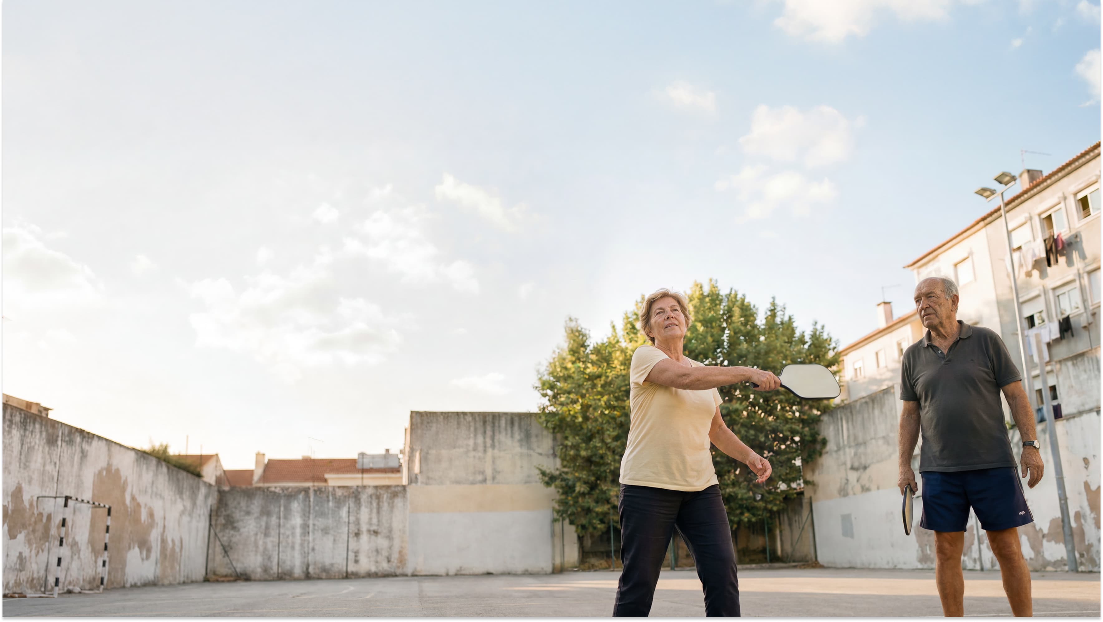 Two older adults play pickleball on an outdoor court. The woman swings a paddle while the man watches. Buildings and a clear sky with clouds are in the background.