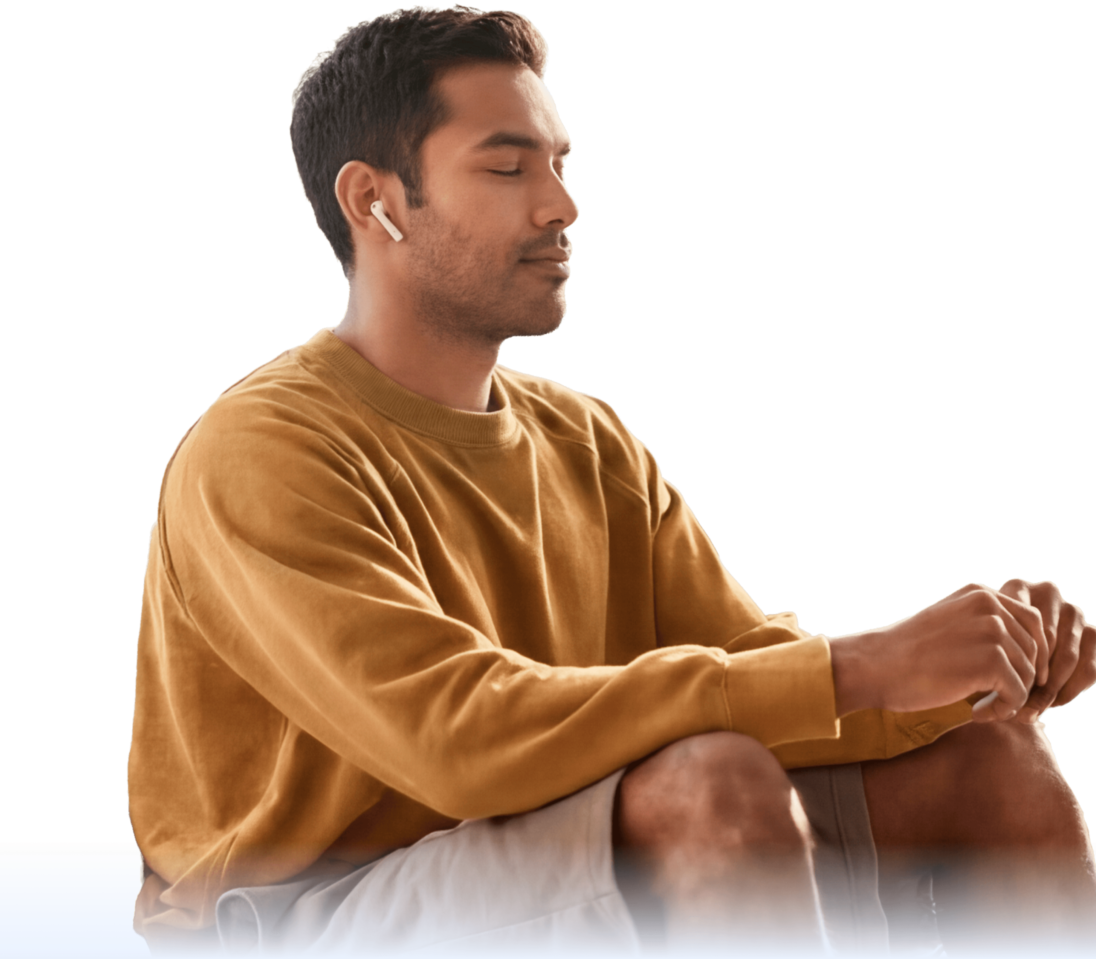 A man wearing a brown sweatshirt and earbuds sits cross-legged with his eyes closed, appearing calm and relaxed against a light background.
