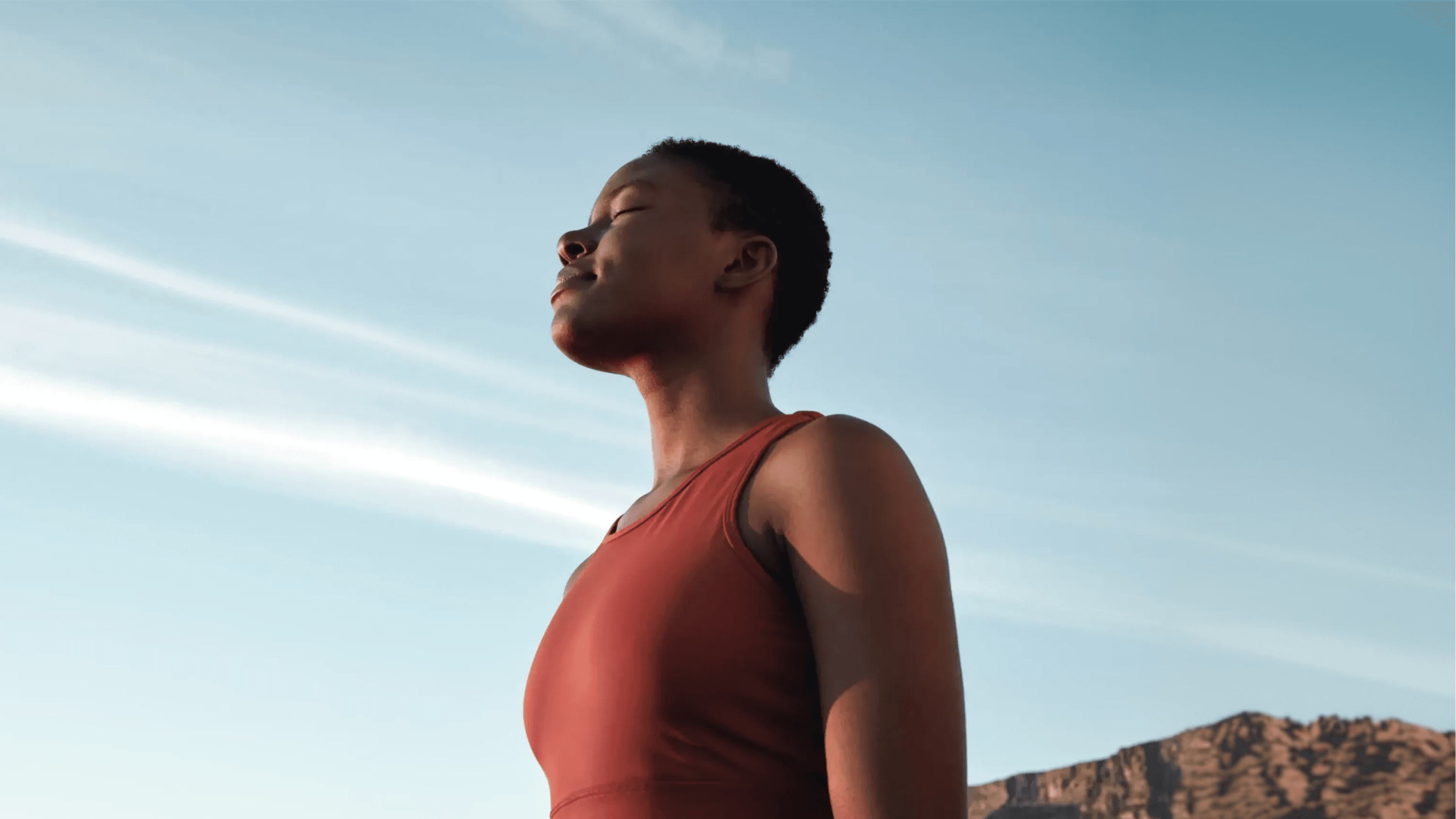 A person in a sleeveless red top stands outdoors with eyes closed, facing the sun, appearing calm and peaceful under a clear blue sky with light clouds and mountains in the background.