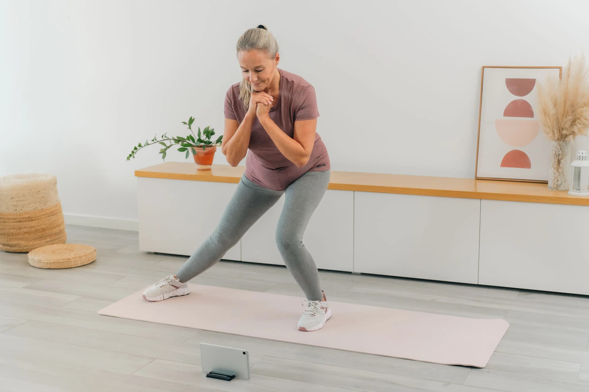 Woman exercising at home, doing a side lunge on a yoga mat, watching a tablet. Room decorated with plants and modern art.
