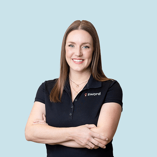 A woman with straight light brown hair, wearing a black collared shirt with a sword logo, stands against a light blue background, smiling with her arms crossed.