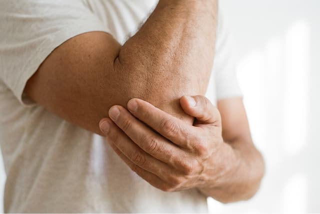 A person wearing a white shirt is holding their elbow with one hand, suggesting discomfort or pain in the elbow area.