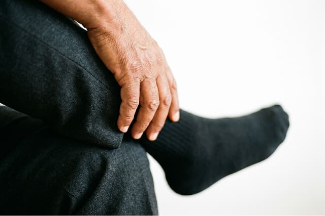 A person wearing dark pants and black socks rests their hand on their knee, with their leg crossed over the other. The background is plain and white.