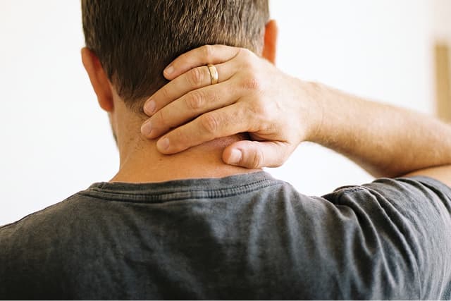 A person seen from behind holds the back of their neck with one hand, suggesting discomfort or pain. The person is wearing a dark shirt and a gold ring on their left hand. The background is out of focus.