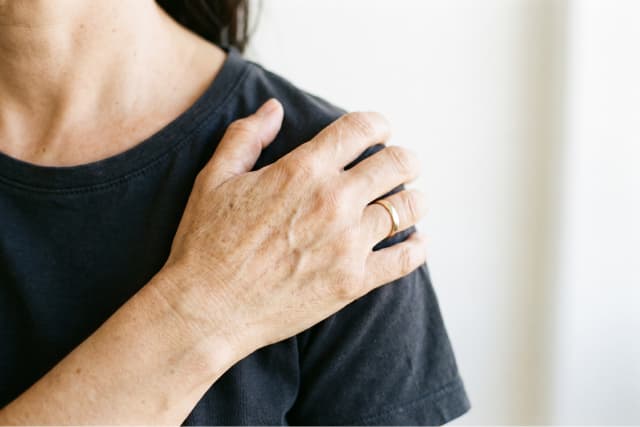 A person wearing a black shirt rests their left hand on their right shoulder, showing a gold ring on their finger. The background is plain and softly lit.