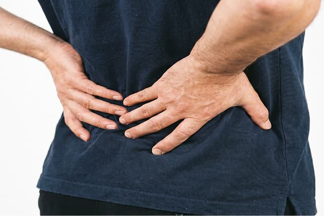 A person wearing a navy blue shirt stands with both hands pressed against their lower back, suggesting discomfort or back pain.