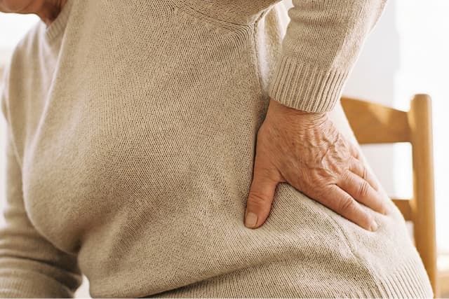An older adult in a beige sweater holds their lower back and hip, showing signs of discomfort or pain, while sitting on a wooden chair indoors.