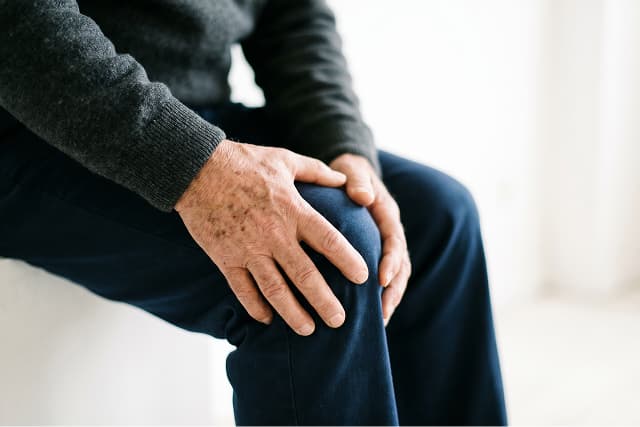 An older adult wearing a dark sweater and pants sits and holds their knee with both hands, appearing to experience knee pain or discomfort.