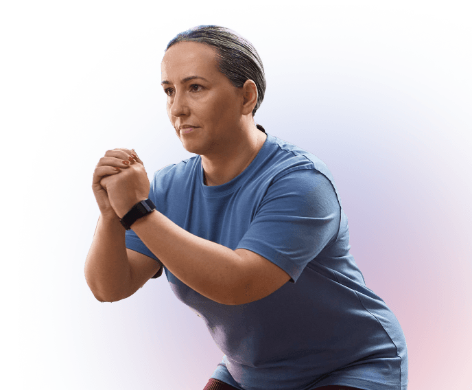 A woman wearing a blue T-shirt and fitness tracker performs a squat exercise, with her hands clasped in front of her chest and a focused expression.