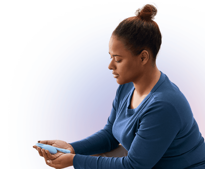 A woman in a blue shirt sits and looks down at a medical device she holds in her hands, appearing focused and thoughtful.