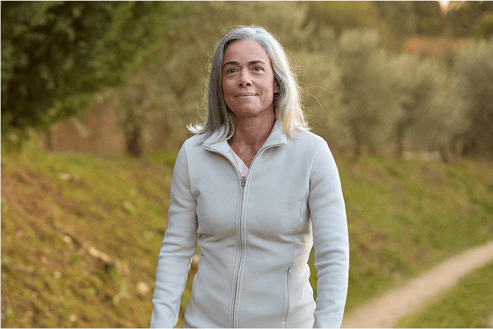 A woman with gray hair walks outdoors on a path, wearing a light-colored zip-up jacket, with greenery in the background.