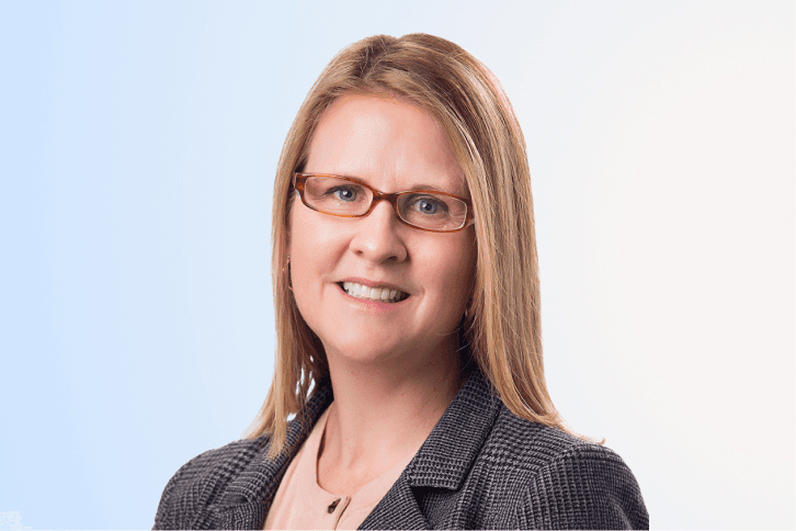A woman with straight, shoulder-length blonde hair wearing glasses, a gray blazer, and a light top, smiles against a light blue and white gradient background.
