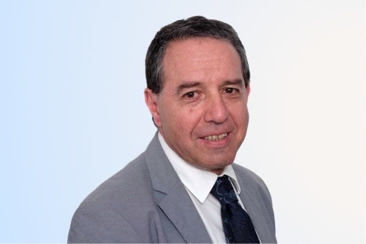 A middle-aged man with short dark hair, wearing a light gray suit, white shirt, and dark patterned tie, smiling slightly while looking at the camera against a light blue and white background.