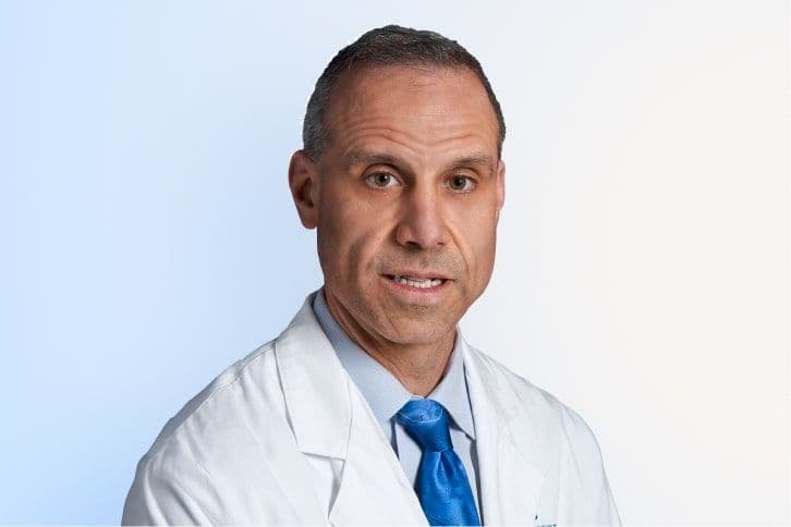 A man wearing a white lab coat, blue dress shirt, and blue tie poses against a light blue and white gradient background. He has short brown hair and a neutral expression.