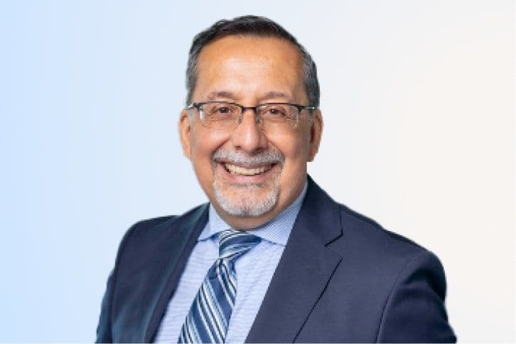 Smiling man with short dark hair, glasses, and a goatee wears a navy suit, blue dress shirt, and striped tie, posed against a light blue and white gradient background.