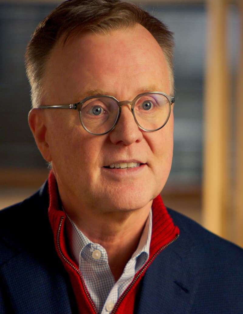 A middle-aged man with short light hair and glasses, wearing a blue blazer, red sweater, and collared shirt, smiles while looking slightly off camera. The background is softly blurred.
