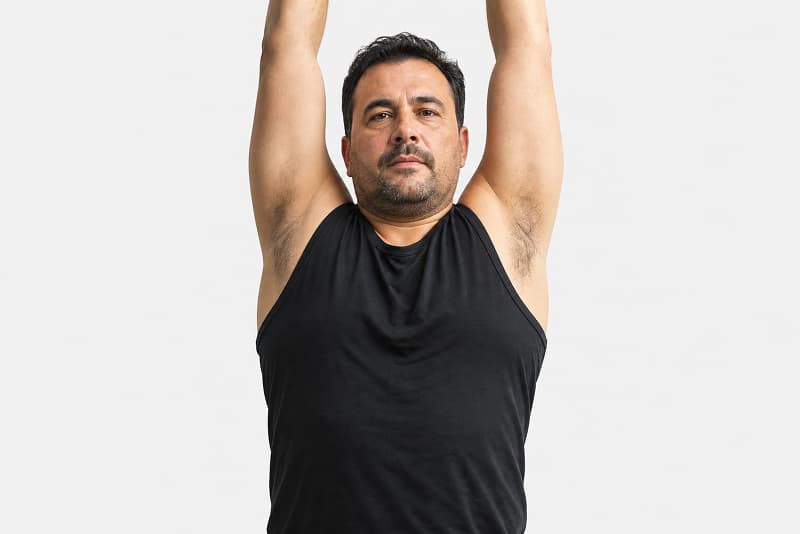 A man wearing a black sleeveless shirt stands with both arms raised overhead, looking straight ahead, against a plain white background.