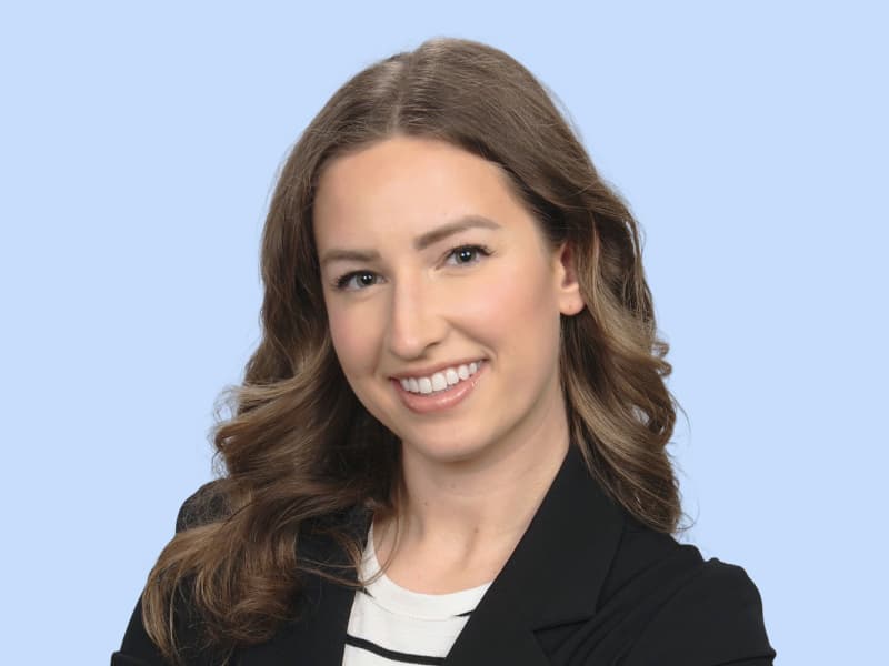 Claire pictured smiling, with long hair in a black blazer and striped shirt, against a light blue background.