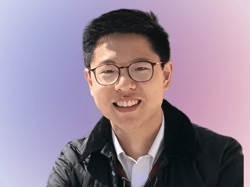 A young person with short dark hair and glasses smiles at the camera, wearing a dark jacket over a white collared shirt, with a soft blue and pink gradient background.